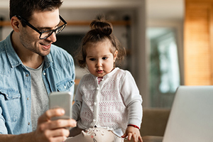 dad working from home inside with his daughter on table showing her phone