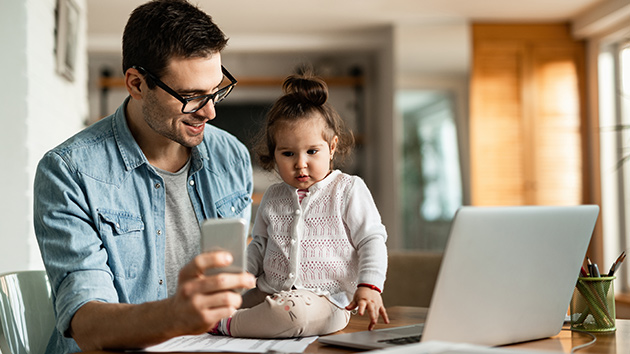 dad working from home inside with his daughter on table showing her phone